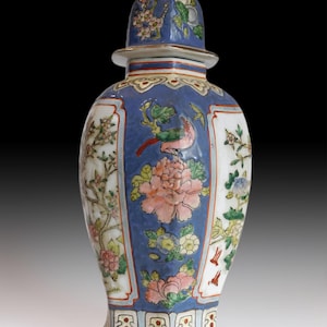 May include: A blue and white ceramic vase with a lid. The vase features floral and bird designs in pink, green, and yellow. The lid and body have a blue background with white panels. The vase is a decorative item.