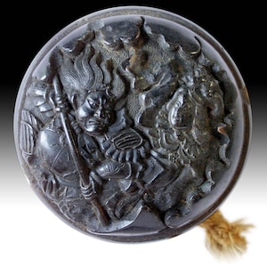 Puede incluir: Un netsuke japonés circular, de color marrón oscuro, con tallas en relieve detalladas. La talla representa dos figuras, una de ellas sosteniendo una lanza, en medio de llamas. Una cuerda texturizada está unida al netsuke.