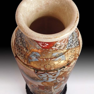 Puede incluir: Un jarrón de cerámica decorativo con una abertura ancha y un diseño detallado y colorido. El jarrón presenta un borde de color crema y una base con intrincados patrones en oro, rojo y azul. Se apoya sobre un soporte negro, mostrando su artesanía ornamentada.