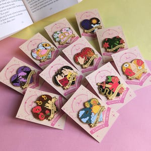 Può includere: Una collezione di spille a forma di cuore in smalto con vari disegni, tra cui fiori, coppie ed elementi celesti. Ogni spilla è su una carta rosa con le parole "Romance" e "Tropes". Un libro aperto è sullo sfondo.