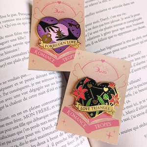 May include: Two heart-shaped enamel pins with gold trim. One features a purple heart with hands reaching for a star, the text "Forbidden Love", and the word "Romance". The other has a black heart with red flowers and the text "Love Triangle".