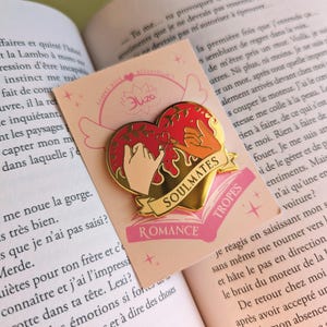 May include: A heart-shaped enamel pin with a gold border, featuring two hands making a pinky promise. The pin has the words "SOULMATES" and "ROMANCE" on it. The pin is on a pink card with the text "Eliza" and a book in the background.