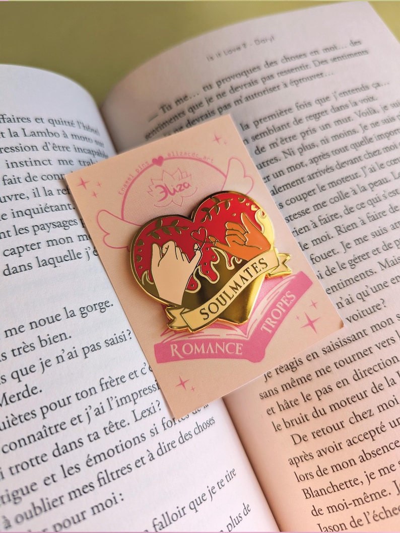 May include: A heart-shaped enamel pin with a gold border, featuring two hands making a pinky promise. The pin has the words "SOULMATES" and "ROMANCE" on it. The pin is on a pink card with the text "Eliza" and a book in the background.