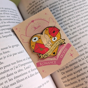 May include: A heart-shaped enamel pin with a gold outline, featuring a hand holding a red envelope and a pair of lips. The pin has a banner that reads "FRIENDS TO LOVERS" and the word "ROMANCE". The pin is on a pink card.