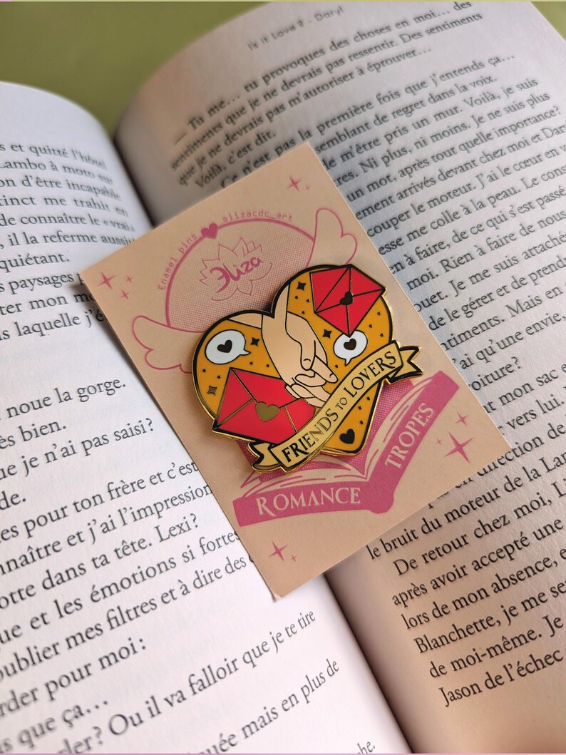 May include: A heart-shaped enamel pin with a gold outline, featuring a hand holding a red envelope and a pair of lips. The pin has a banner that reads "FRIENDS TO LOVERS" and the word "ROMANCE". The pin is on a pink card.
