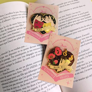 May include: Two heart-shaped enamel pins with gold accents. One features theater masks and the text "FAKE DATING", the other has flowers and the text "SLOW BURN". Both pins have the words "ROMANCE" and "TROPES".