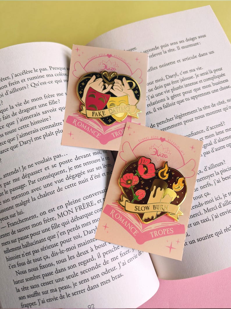 May include: Two heart-shaped enamel pins with gold accents. One features theater masks and the text "FAKE DATING", the other has flowers and the text "SLOW BURN". Both pins have the words "ROMANCE" and "TROPES".