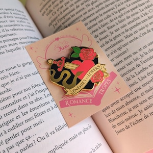 May include: A gold-colored enamel pin with a heart pierced by a sword, surrounded by roses and a black snake. The pin has the words "Enemies to Lovers" and "Tropes" on it. The pin is on a pink card.