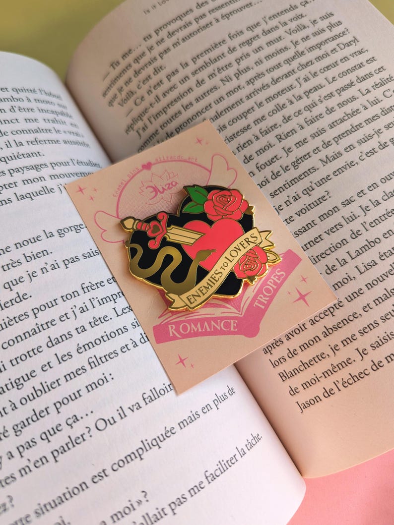 May include: A gold-colored enamel pin with a heart pierced by a sword, surrounded by roses and a black snake. The pin has the words "Enemies to Lovers" and "Tropes" on it. The pin is on a pink card.