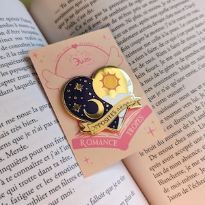 May include: A heart-shaped enamel pin with a celestial and sun design. The pin features a navy blue night sky with stars and a crescent moon, and a golden sun with clouds. The banner reads "Opposites Attract Romance Tropes."