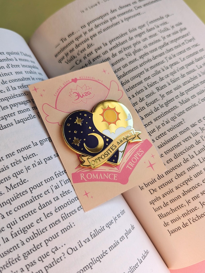 May include: A heart-shaped enamel pin with a celestial and sun design. The pin features a navy blue night sky with stars and a crescent moon, and a golden sun with clouds. The banner reads "Opposites Attract Romance Tropes."