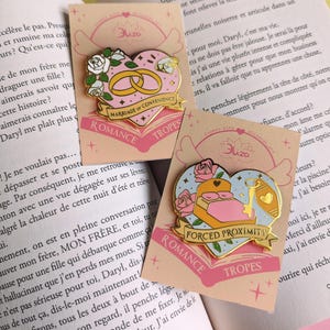 May include: Two enamel pins on cards with a book background. The pins are heart-shaped with gold accents. One features wedding rings and the text "Marriage of Convenience." The other shows a bed and key with the text "Forced Proximity."