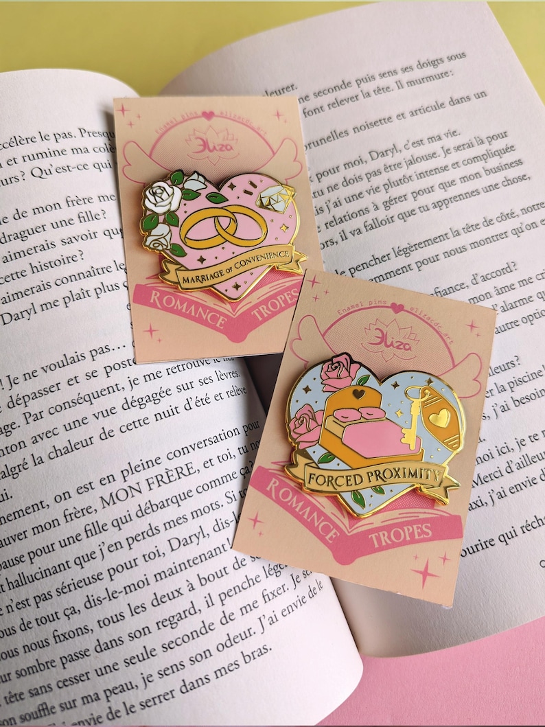 May include: Two enamel pins on cards with a book background. The pins are heart-shaped with gold accents. One features wedding rings and the text "Marriage of Convenience." The other shows a bed and key with the text "Forced Proximity."