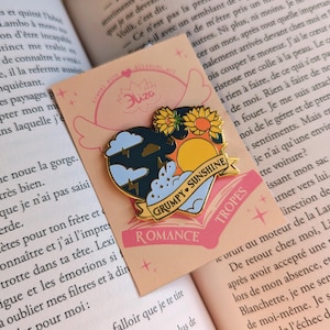 May include: Enamel pin with a heart-shaped design featuring a stormy cloud, a sunflower, and a sunny sky. The pin includes the text "GRUMPY SUNSHINE" and "ROMANCE" on a banner. The pin is on a peach-colored card.