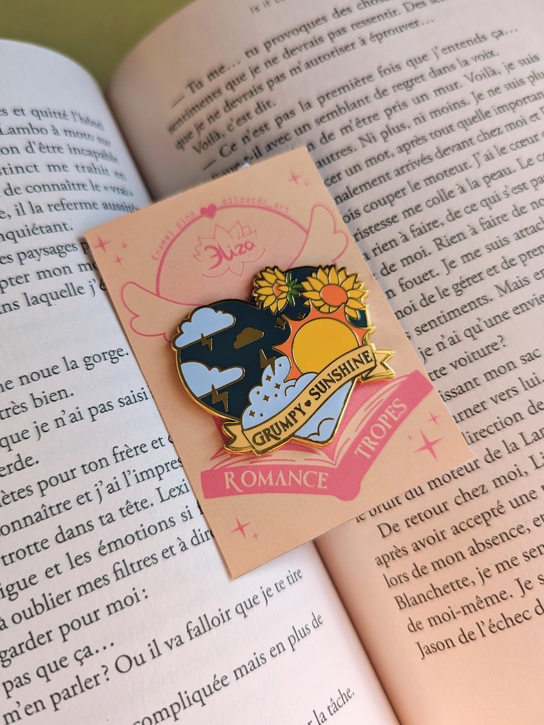 May include: Enamel pin with a heart-shaped design featuring a stormy cloud, a sunflower, and a sunny sky. The pin includes the text "GRUMPY SUNSHINE" and "ROMANCE" on a banner. The pin is on a peach-colored card.