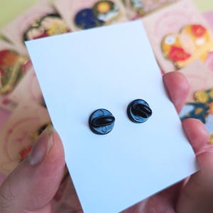May include: Close-up of two black enamel pin backs on a white card, held by a person. The pin backs are round with a small post and clasp. Other colorful pin cards are visible in the background.