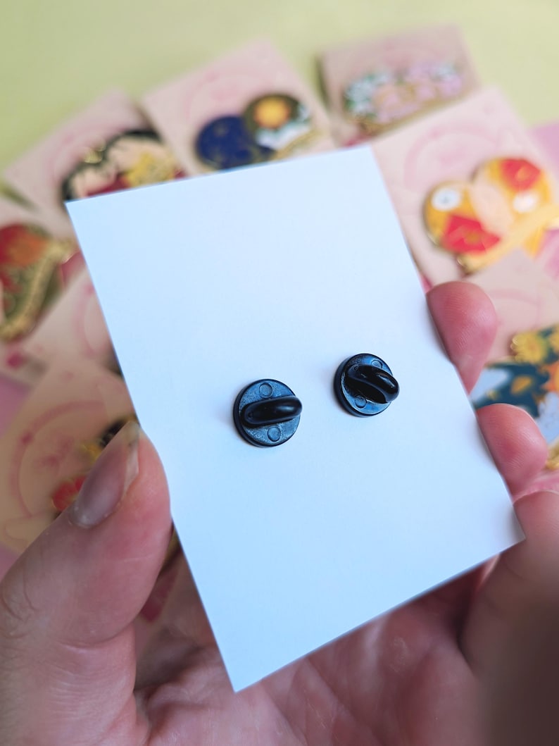 May include: Close-up of two black enamel pin backs on a white card, held by a person. The pin backs are round with a small post and clasp. Other colorful pin cards are visible in the background.