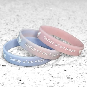 May include: Three silicone wristbands with the text "Daddy of an Angel" in white lettering. The wristbands are in shades of blue and pink.