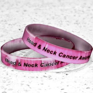May include: Two pink silicone wristbands with the text "Head & Neck Cancer Awareness" in black. The bands are stacked on top of each other, with a marbled effect on the surface.