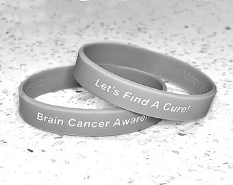 May include: Two gray silicone wristbands. One reads "Brain Cancer Awareness" and the other says "Let's Find A Cure!" with the website address and brand name printed on the bands. The bands are stacked on top of each other.