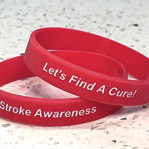 Peut inclure: Deux bracelets de poignet rouges en silicone. L'un porte l'inscription "Stroke Awareness" et l'autre "Let's Find A Cure !" en lettres majuscules blanches. Les bracelets sont empilés l'un sur l'autre.