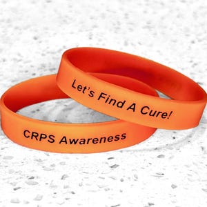 May include: Two orange silicone wristbands with black text. One band reads "CRPS Awareness," and the other says "Let's Find A Cure!" The bands are stacked on top of each other.