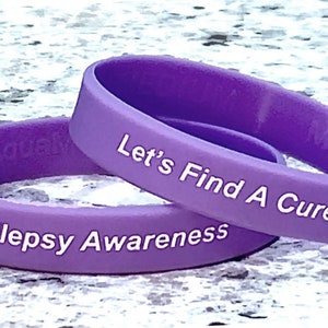 May include: Two purple silicone wristbands with white lettering. The top band reads "Let's Find A Cure!" and the bottom band reads "Epilepsy Awareness".