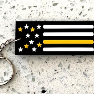 May include: Black and white American flag keychain with a yellow stripe, representing support for law enforcement. The keychain has a silver chain and ring.