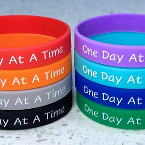 May include: A stack of eight silicone wristbands in various colors, each with the text "One Day At A Time" printed in white.