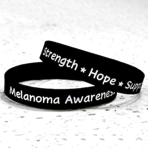May include: Two black rubber bracelets with white text. One bracelet is lying flat, and the other is partially on top. The text on the bracelets reads "Strength * Hope * Support" and "Melanoma Awareness".