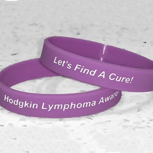 May include: Two purple silicone wristbands with white text. The top band reads "Let's Find A Cure!" and the bottom band reads "Hodgkin Lymphoma Awareness".