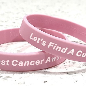 May include: Two pink silicone bracelets with white text. One reads "Breast Cancer Awareness" and the other says "Let's Find A Cure!". The bracelets are stacked on a white speckled surface.