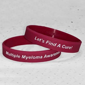 May include: Two red silicone wristbands with white text. The top band reads "Let's Find A Cure!" and the bottom band reads "Multiple Myeloma Awareness".