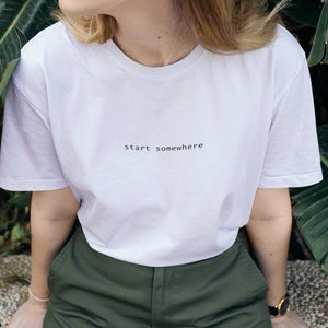 May include: A white t-shirt with the phrase "start somewhere" printed in black. The shirt is paired with olive green pants. The model is wearing a gold watch with a black band.