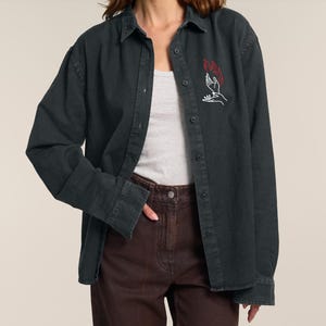 Può includere: Una camicia di jeans grigio scuro con un disegno ricamato bianco e rosso di due mani che tengono delle fiamme.