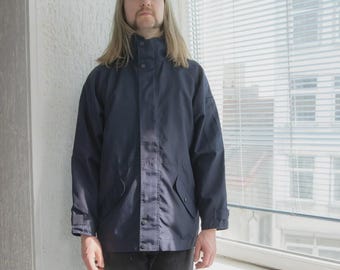 Pode incluir: Uma jaqueta azul marinho com gola alta e fecho de botão. A jaqueta tem dois bolsos frontais e é usada com jeans pretos e botas pretas. A pessoa está em frente a uma janela com persianas.
