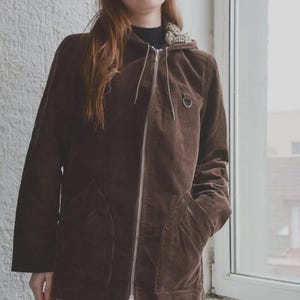 Może przedstawiać: Brązowa kurtka sztruksowa z kapturem i srebrnym zamkiem błyskawicznym. Kurtka ma dwie przednie kieszenie i kaptur ze sznurkiem. Modelka ma na sobie czarny top i kremowe spodnie.