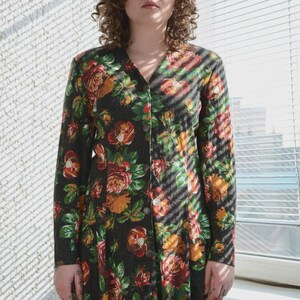 May include: A long-sleeved, button-down dress with a floral pattern in shades of red, orange, and green on a dark background. The dress has a V-neck and is worn by a person standing near a window with blinds.