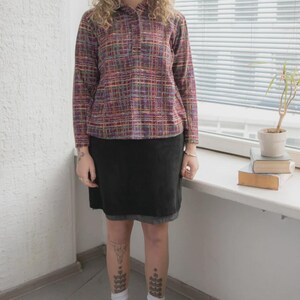 May include: A person wearing a long-sleeved, button-down shirt with a plaid pattern in shades of purple, red, and yellow. The person is also wearing a black skirt, white socks, and red shoes. The outfit is casual and stylish.