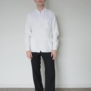 May include: A person wearing a white long-sleeved shirt with a mandarin collar and a full-length zip, paired with black trousers and black shoes. The outfit is simple and classic.