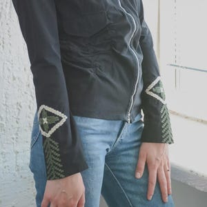 Puede incluir: Chaqueta negra con cremallera blanca y bordados decorativos en las mangas. Las mangas presentan un diseño geométrico verde y blanco. La chaqueta se combina con vaqueros azules.