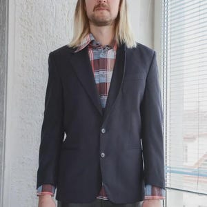 Op de afbeelding: Een marineblauwe blazer met twee knopen, gedragen over een geruit overhemd. De blazer heeft een klassiek ontwerp en wordt gecombineerd met een donkere broek. De outfit is geschikt voor een business casual of smart casual look.