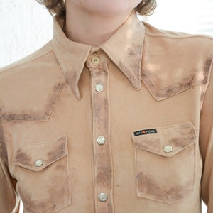 K&ouml;nnte beinhalten: Ein hellbraunes Wildledershirt mit Knopfleiste und einer einzigen Brusttasche. Das Shirt hat einen verblassten, Vintage-Look und ein kleines schwarzes Patch mit dem Text "ENERGIE" auf der linken Brustseite.