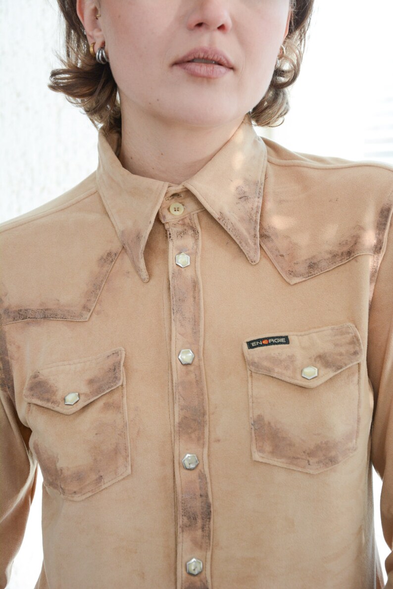 K&ouml;nnte beinhalten: Ein hellbraunes Wildledershirt mit Knopfleiste und einer einzigen Brusttasche. Das Shirt hat einen verblassten, Vintage-Look und ein kleines schwarzes Patch mit dem Text "ENERGIE" auf der linken Brustseite.