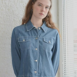 May include: A light blue denim jacket with a button-up front, two chest pockets, and a collared neckline. The jacket is paired with a bright pink skirt. The model is standing in front of a window with blinds.