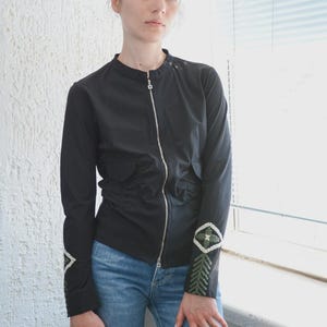 Puede incluir: Chaqueta negra con cremallera, cuello alzado y bordados decorativos en las mangas. La chaqueta tiene una silueta entallada y se combina con vaqueros azules. Las mangas presentan patrones geométricos verdes y blancos.