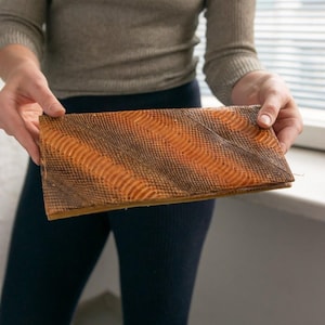 Könnte beinhalten: Rechteckige Clutch mit strukturierter Oberfläche und Muster. Sie weist diagonale Bänder in Orange und Braun auf, die einen einzigartigen visuellen Effekt erzeugen. Das Design deutet auf eine Vintage- oder handwerkliche Ästhetik hin.