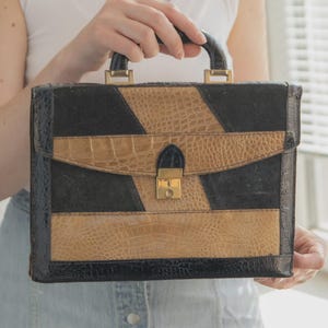May include: A vintage briefcase with a black and tan faux crocodile skin pattern. The briefcase has a gold-colored clasp and handle hardware. The bag is rectangular and has a flap closure.