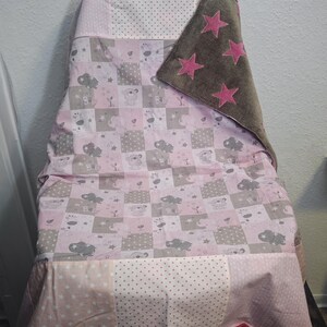 May include: A baby blanket draped over a chair, featuring a patchwork design with pink, gray, and white squares. The blanket has a soft, plush brown section with pink star appliqu&eacute;s. The design includes animal and cloud motifs.