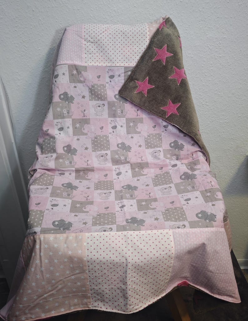 May include: A baby blanket draped over a chair, featuring a patchwork design with pink, gray, and white squares. The blanket has a soft, plush brown section with pink star appliqu&eacute;s. The design includes animal and cloud motifs.